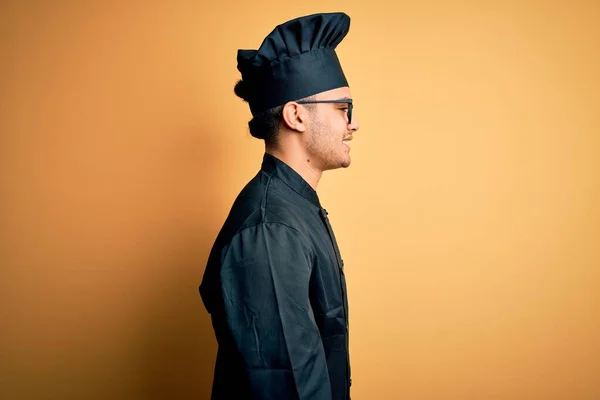 Young brazilian chef man wearing cooker uniform and hat over isolated yellow background looking to side, relax profile pose with natural face and confident smile.