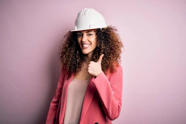 年轻美丽的建筑师女人 卷曲的头发 戴着安全帽 粉红的背景 手举着快乐的大拇指 在镜头前看着成功的表情 — 图库照片