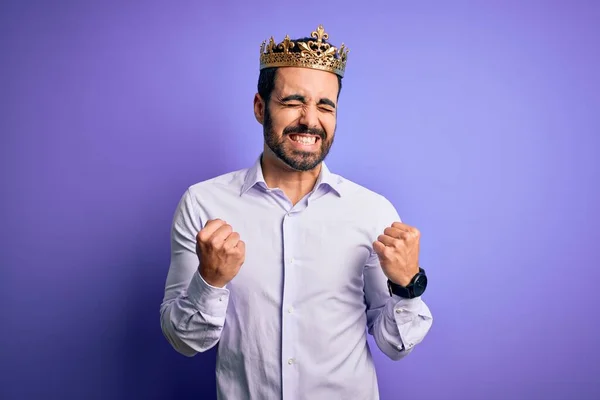 年轻英俊的大胡子男子 头戴金色王冠 身披紫色背景 为成功而兴奋 举起双臂 闭上双眼 微笑着庆祝胜利 赢的概念 — 图库照片