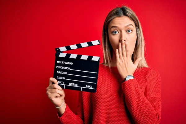 Young beautiful woman holding clapboard standing over isolated red background cover mouth with hand shocked with shame for mistake, expression of fear, scared in silence, secret concept