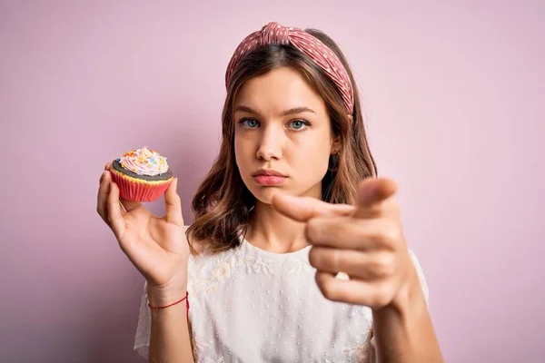 若いですブロンド女の子食べるA甘いですチョコレートカップケーキ以上ピンク隔離された背景Pointingに指でカメラとあなたに 手のサイン 正と自信のジェスチャーからザフロント — ストック写真