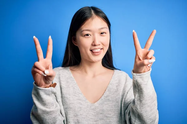 青い隔離された背景の上に立つカジュアルなセーターを着た若い美しいアジアの女性は 勝利のサインをして両手の指を見せて舌で笑っています 第二番 — ストック写真