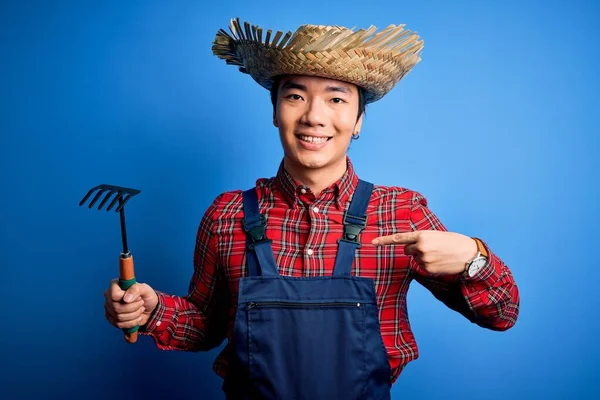 Young handsome chinese farmer man wearing apron and straw hat holding rake tool with surprise face pointing finger to himself