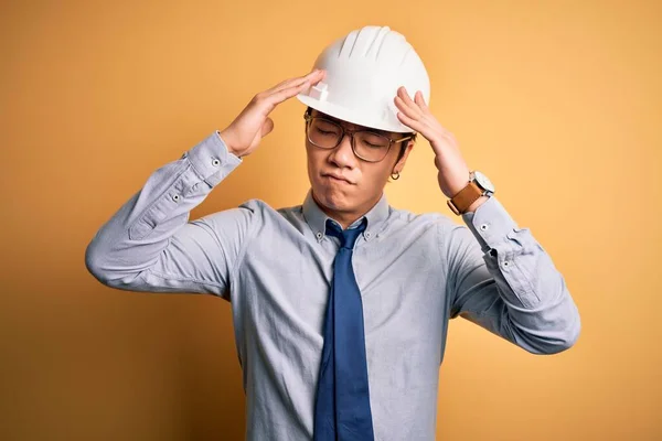 若いハンサムな中国の建築家の男性は 安全ヘルメットを着用し 痛みや片頭痛のために必死の頭痛やストレスに苦しんで黄色の背景にネクタイ 手を頭に — ストック写真