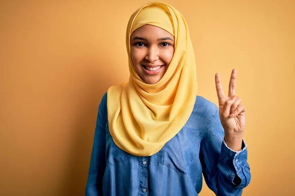黄色の背景にアラブの伝統的なヒジャーブを身に着けている巻き毛を持つ若い美しい女性は 勝利のサインを行う指を示すカメラを見て笑っています 第二番 — ストック写真