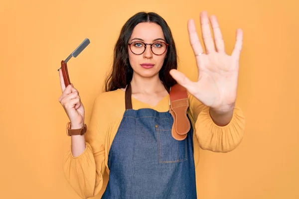 Young beautiful barber woman wearing apron holding razor blade over yellow background with open hand doing stop sign with serious and confident expression, defense gesture