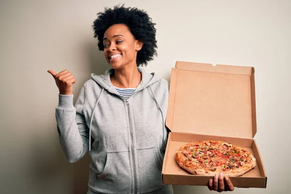 Young African American afro woman with curly hair holding delivery box with Italian pizza pointing and showing with thumb up to the side with happy face smiling