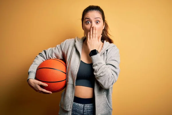 若い美しいブルネットのスポーツウーマンは 黄色の背景カバー口の上にバスケットボールボールを保持します手でショックを受けました恥でミス 恐怖の表現 沈黙の中で怖がって 秘密の概念 — ストック写真