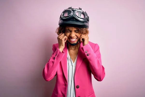 非洲裔美国骑摩托车的女人 卷曲的头发 粉色背景下头戴安全帽 用手指捂住耳朵 对嘈杂的音乐感到恼怒 聋人概念 — 图库照片
