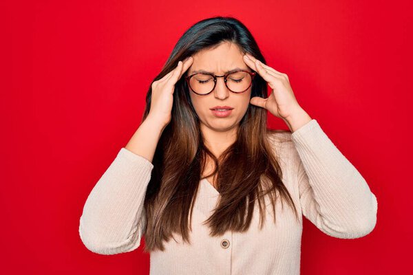 Young hispanic smart woman wearing glasses standing over red isolated background with hand on head for pain in head because stress. Suffering migraine.