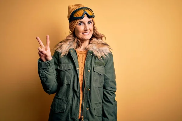 中年漂亮的金发滑雪者女人穿着雪地运动鞋和滑雪护目镜 带着自信而快乐的微笑 同时又用手指指了指二号 — 图库照片