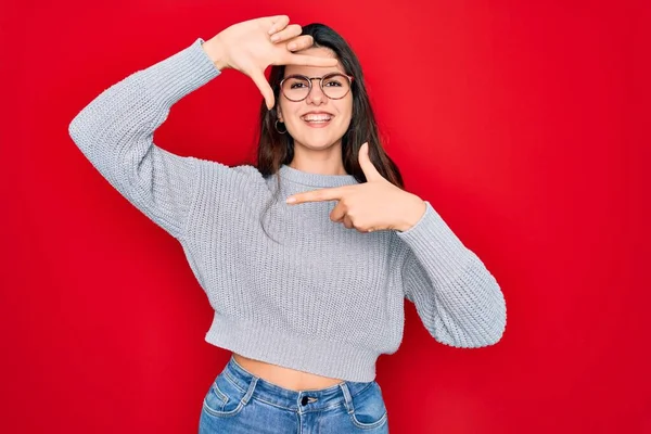 若い美しいブルネットの女性は幸せな顔で手と指でフレームを作る笑顔赤い背景の上にカジュアルなセーターを着ています 創造性と写真の概念 — ストック写真