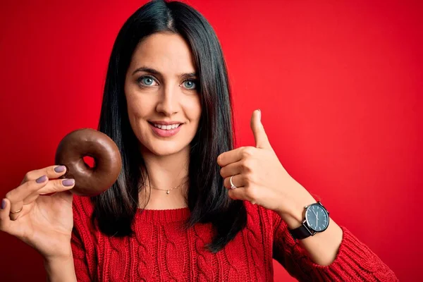 年轻的黑发女人 蓝眼睛 吃巧克力甜甜圈而不是红的孤立的背景 开心的笑着做个好手势 用手指竖起大拇指 很好的标志 — 图库照片