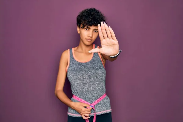Beautiful african american sporty woman doing exercise controlling weight using tape measure with open hand doing stop sign with serious and confident expression, defense gesture