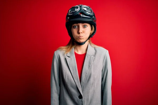 若い美しいブロンドのオートバイの女性は 面白い顔で頬をむき出す赤い背景にオートバイのヘルメットをかぶっています 口は空気で膨脹し 狂気の表現 — ストック写真
