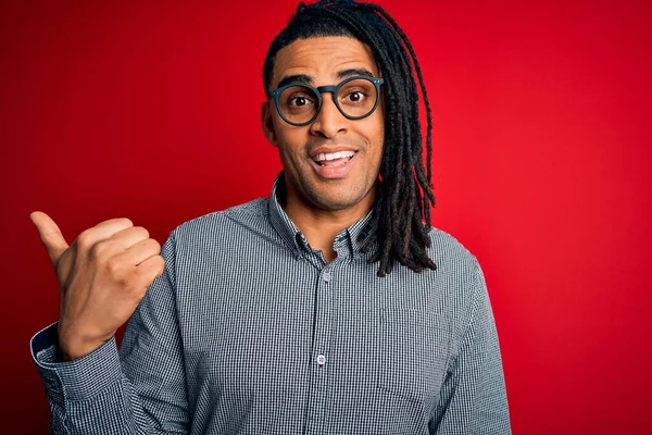 Young handsome african american man with dreadlocks wearing casual shirt and glasses smiling with happy face looking and pointing to the side with thumb up.