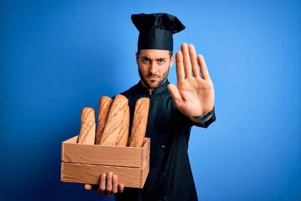 Young cooker man with beard wearing uniform holding box with bread over blue background with open hand doing stop sign with serious and confident expression, defense gesture