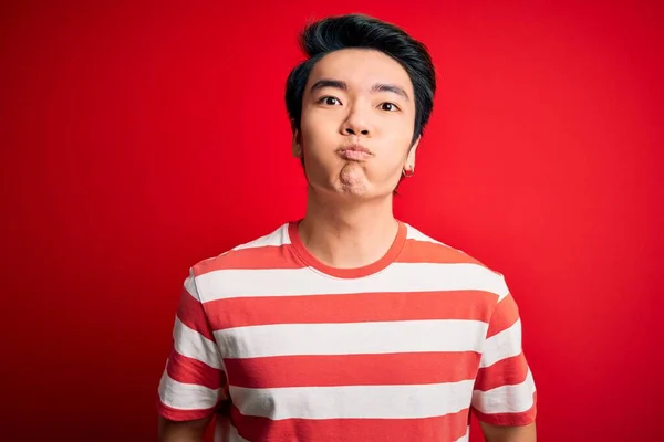 若いハンサムな中国人男性が面白い顔で頬をむち打つ赤い背景の上に立ってカジュアルな縞模様のTシャツを着て 口は空気で膨脹し 狂気の表現 — ストック写真