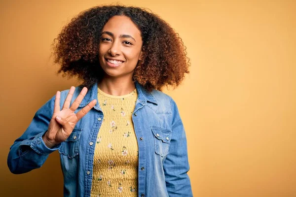 若いですアフリカ系アメリカ人女性とともにアフロ髪身に着けていますカジュアルなデニムシャツ上の黄色の背景ショーや指の数4で指摘しながら 自信と幸せ — ストック写真