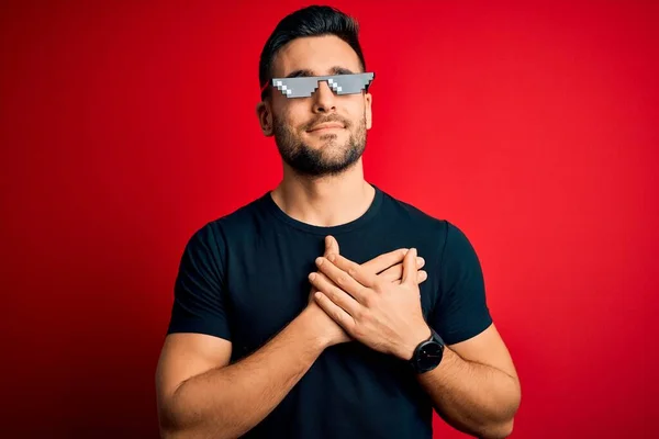 Young handsome man wearing funny thug life sunglasses over isolated red background smiling with hands on chest with closed eyes and grateful gesture on face. Health concept.