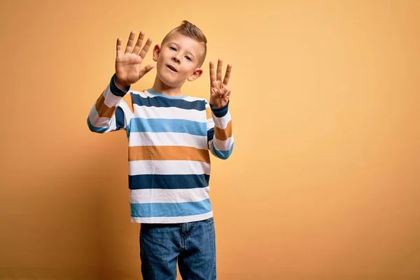 黄色の背景にカラフルなストライプのシャツを身に着けている青い目をした若い白人の子供は自信を持って幸せな笑みを浮かべている間 指の数8で示すと指摘 — ストック写真