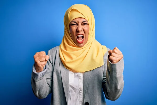 中年黑发女商人身穿穆斯林传统的头巾 头戴蓝色背景的头巾 怒气冲冲 怒气冲冲地举起拳头 一边大喊大叫 愤怒和好斗的概念 — 图库照片