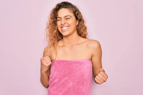 Beautiful blonde woman with blue eyes wearing towel shower after bath over pink background excited for success with arms raised and eyes closed celebrating victory smiling. Winner concept.