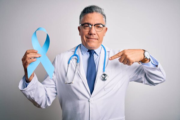 Мужчина средних лет с седыми волосами, в пальто, с голубой раковой лентой, с удивленным лицом, указывающим на себя пальцем.
