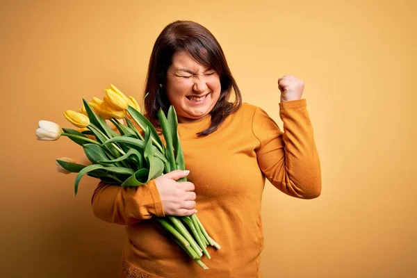 Beautiful plus size woman holding romantic bouquet of natural tulips flowers over yellow background very happy and excited doing winner gesture with arms raised, smiling and screaming for success. Celebration concept. — Stock Image Beautiful Size Woman Holding Romantic Bouquet Natural Tulips Flowers Yellow Stock Photo