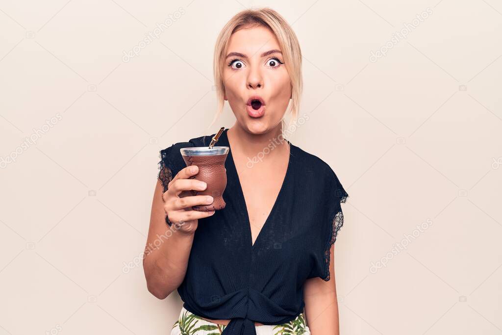 Young beautiful blonde woman drinking cup of mate infusion beverage over white background scared and amazed with open mouth for surprise, disbelief face