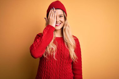 Genç, güzel, sarışın bir kadın. Günlük kazak ve beyaz arka planda yün şapka takıyor. Bir gözünü eliyle kapatıyor..