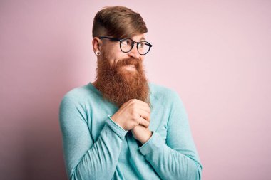 Pembe arka planda gözlük takan, sakallı yakışıklı kızıl saçlı, gergin ve heyecanlı bir şekilde çenesi yan tarafa bakıyor.