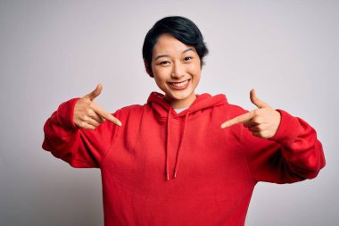 Genç, güzel Asyalı bir kız beyaz arka planda kapüşonlu sıradan bir sweatshirt giyiyor. Kendine güvenen, yüzünde gülümseme olan, kendini gururlu ve mutlu gösteren bir kız..