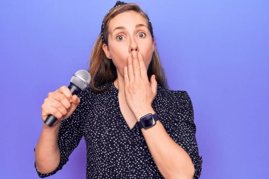 Mor arka planda şarkı söyleyen, ağzı elle kaplayan, şok olmuş ve hata yapmaktan korkan güzel sarışın bir kadın. şaşırmış ifade 