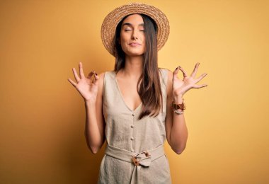 Genç ve güzel esmer kadın tatilde. Günlük elbiseler ve şapkalar giyiyor. Gözleri kapalı rahatlıyor ve parmaklarıyla meditasyon hareketi yapıyor. Yoga konsepti.