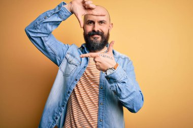Sakallı, sıradan bir kot ceket ve çizgili tişört giyen, elleri ve parmakları mutlu yüzüyle çerçeve yapan yakışıklı kel adam. Yaratıcılık ve fotoğrafçılık kavramı.