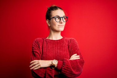 Genç, güzel, kızıl saçlı bir kadın izole edilmiş kırmızı arka plan üzerine günlük süveter giyiyor. Kollarını kavuşturmuş, ikna olmuş ve kendinden emin bir şekilde yan tarafa bakıyor.