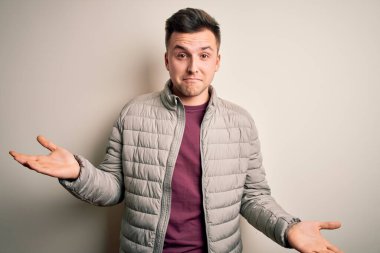 Sıradan bir kışlık ceket giyen yakışıklı beyaz adam izole arka planda duruyor. Kolları ve elleri havada, kafası karışmış bir ifade takınıyor. Şüphe kavramı.