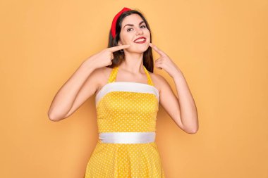 Sarı arka planda 50 'li yılların moda elbisesini giyen, neşeli gülümseyen ve parmaklarıyla ve ağzıyla işaret eden genç ve güzel bir kadın. Diş sağlığı konsepti.