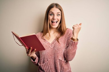 Genç, güzel, kızıl saçlı, kitap okuyan, izole edilmiş beyaz arka planda gururla bağıran, zaferi ve başarıyı kutlayan çok heyecanlı, coşkulu, neşeli bir kadın.