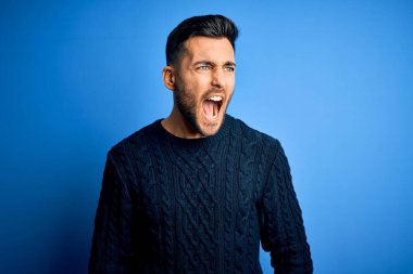 İzole edilmiş mavi arka planda dikilen sıradan bir süveter giyen genç yakışıklı sinirli ve öfkeli çığlıklar atan, öfkeyle bağıran. Öfke ve agresif kavram.