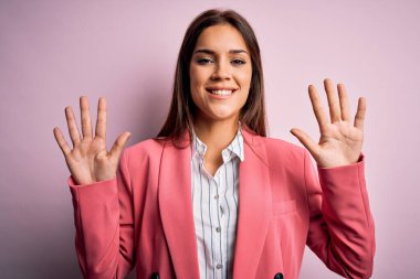 Güzel, esmer, ceketli bir kadın pembe arka planda dikiliyor. 10 numaralı parmaklarıyla işaret ediyor. Kendinden emin ve mutlu bir şekilde gülümsüyor..