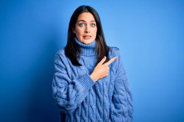 Mavi gözlü, balıkçı yaka kazak giyen genç esmer kadın endişeli ve gergin bir kenara işaret ediyor.