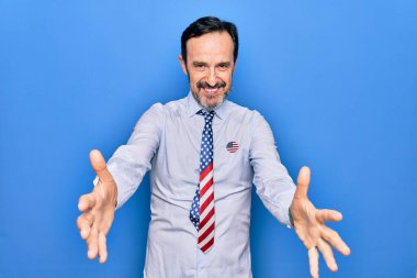 Orta yaşlı, vatansever bir iş adamı mavi arka plan üzerinde kravat takıyor ve kameraya sarılıp gülümsüyor. Mutluluğu kucaklayan neşeli bir ifade..