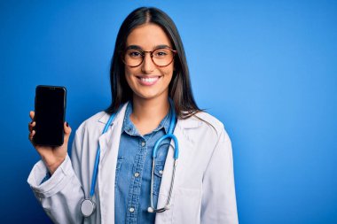 Genç, güzel, esmer, gözlüklü ve ceketli bir kadın elinde mutlu bir yüzle ayakta duran ve gülümseyen bir gülümsemeyle dişlerini gösteren akıllı bir telefon tutuyor.