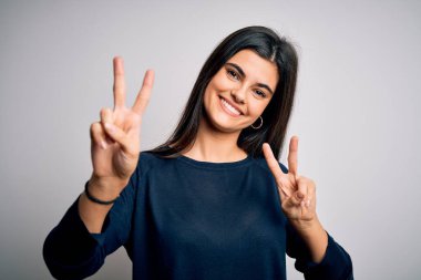 Genç, güzel, esmer, sıradan bir süveter giyen, beyaz arka planda durup gülümseyen, parmaklarını zafer işareti yaparken gösteren kameraya bakan bir kadın. İki numara..