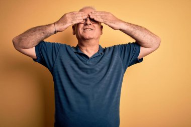 Orta yaşlı yakışıklı beyaz bir adam sarı arka plan üzerine günlük polo ve gözlük takıyor. Gözleri güler yüzlü ve komik ellerle örtüyor. Kör kavram.