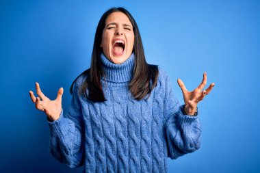 Mavi gözlü genç esmer kadın balıkçı yaka kazak giyiyor deli deli deli bağırıyor ve agresif bir ifade ve kollar havada bağırıyor. Hayal kırıklığı konsepti.