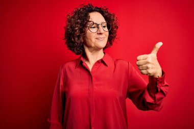 Orta yaşlı güzel kıvırcık saçlı kadın kırmızı arka planda günlük gömlek ve gözlük takıyor. Gururlu görünüyor, gülümsüyor.