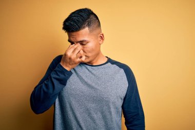 Sarı arka planda dikilen gündelik tişört giyen genç, yakışıklı, latin adam yorgun, burnu ve gözleri ovuşturup yorgun ve baş ağrısı çekiyor. Stres ve hüsran kavramı.